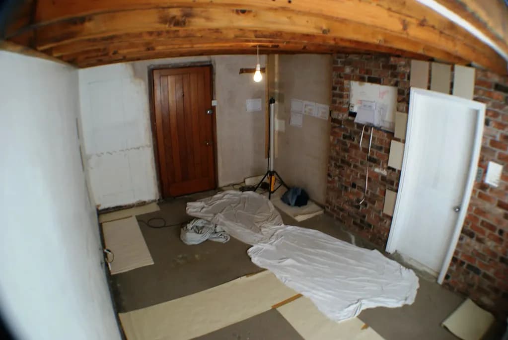 A bare room before fit-out, with dust sheets on the floor, exposed timber ceiling, brick wall and a work light on a tripod