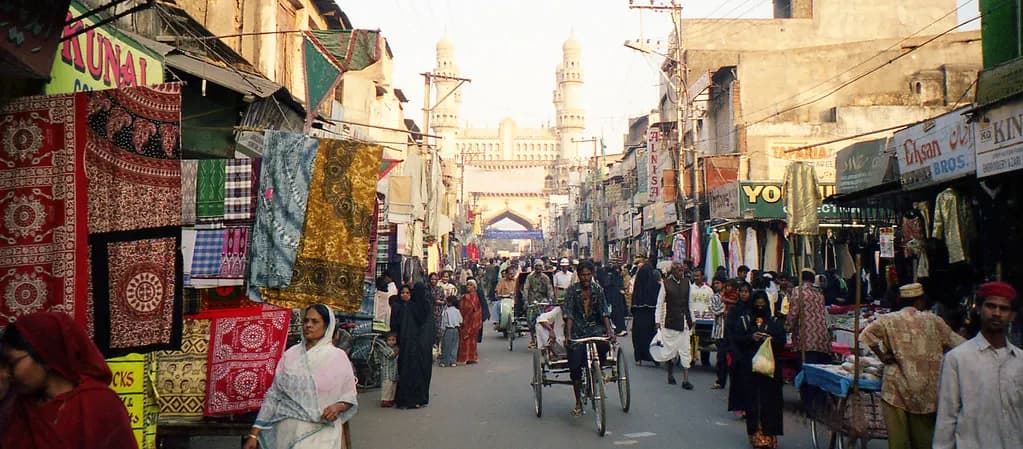 A crowded shopping street in Hyderabad's old city with textile shops and the Charminar in the background
