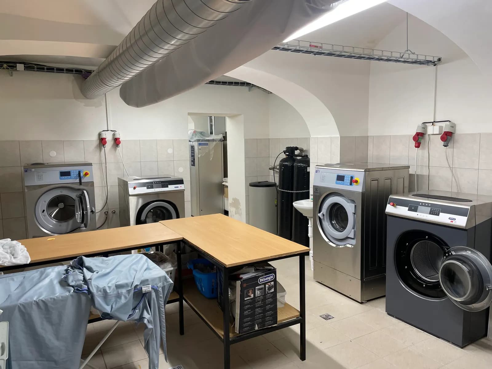 A small commercial laundry room with front-loading washer-extractors, tumble dryers, a folding table and ceiling ducting