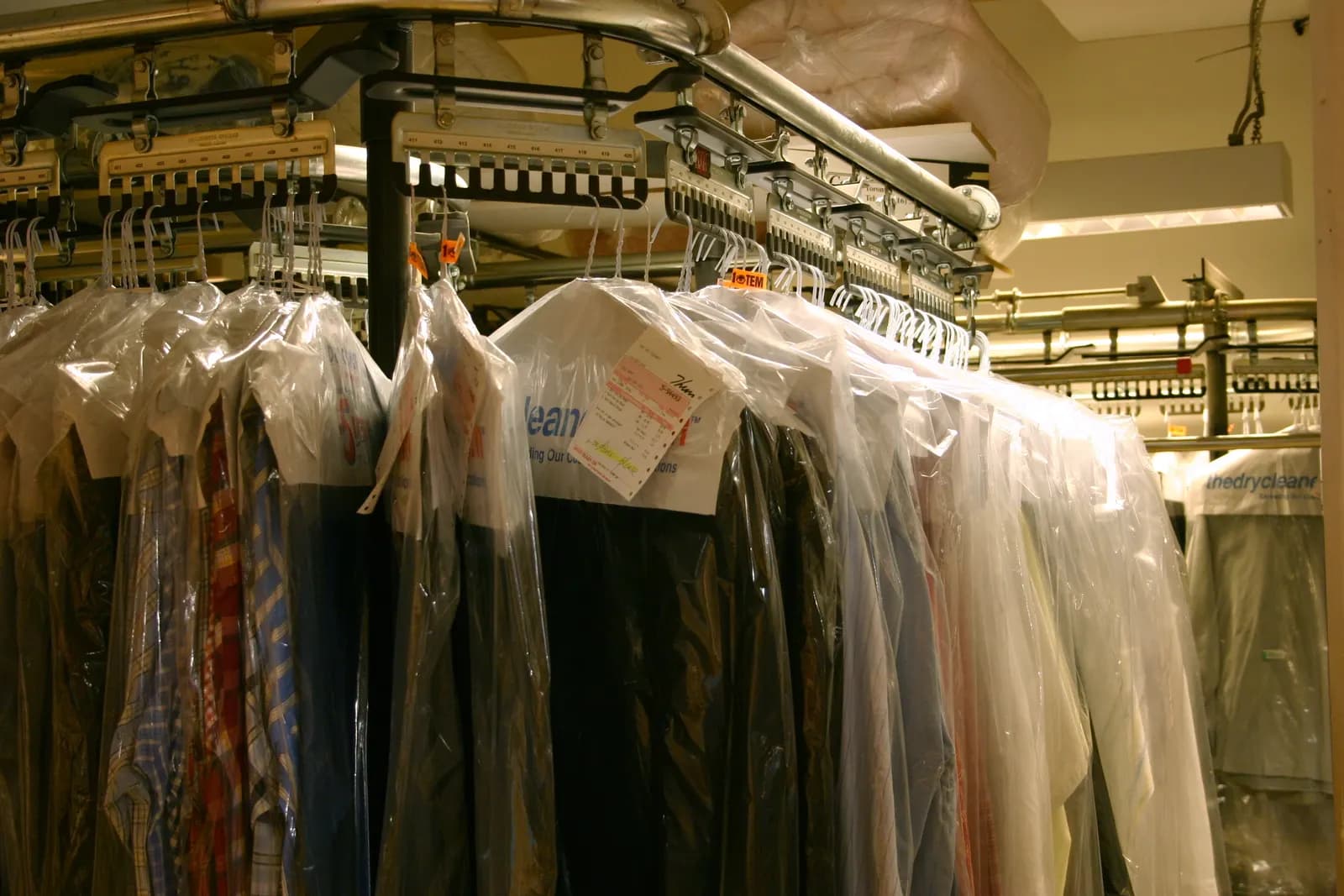 Rows of dry-cleaned garments in clear plastic covers hanging on a powered conveyor rack with paper tags