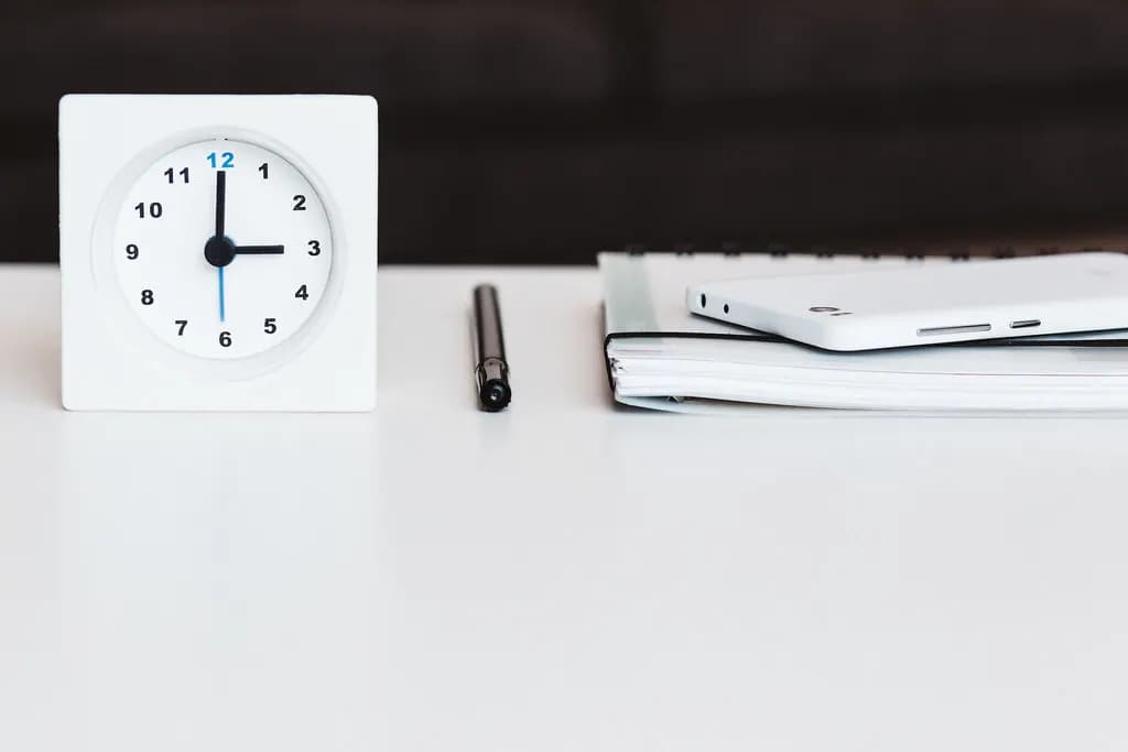 A white desk clock, pen and notebook on an empty office desk