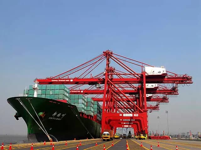 A container ship being worked by red gantry cranes at a container terminal at Jawaharlal Nehru Port, Navi Mumbai, with trucks waiting on the quay