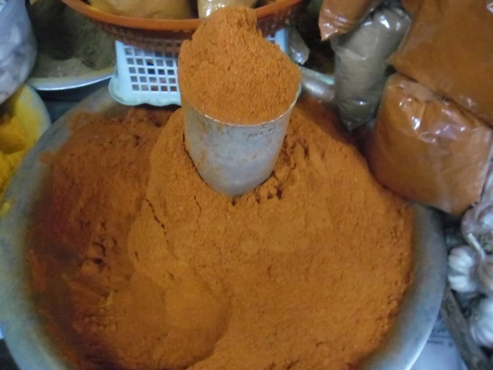 A heaped steel bowl of loose red chilli powder with a measuring cup, among other spice powders at a market stall