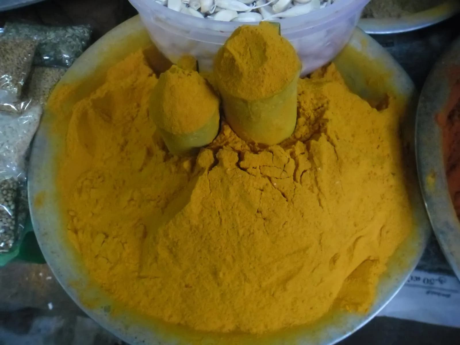 Loose turmeric powder piled in a wide steel bowl at a market stall, scooped with a steel tumbler