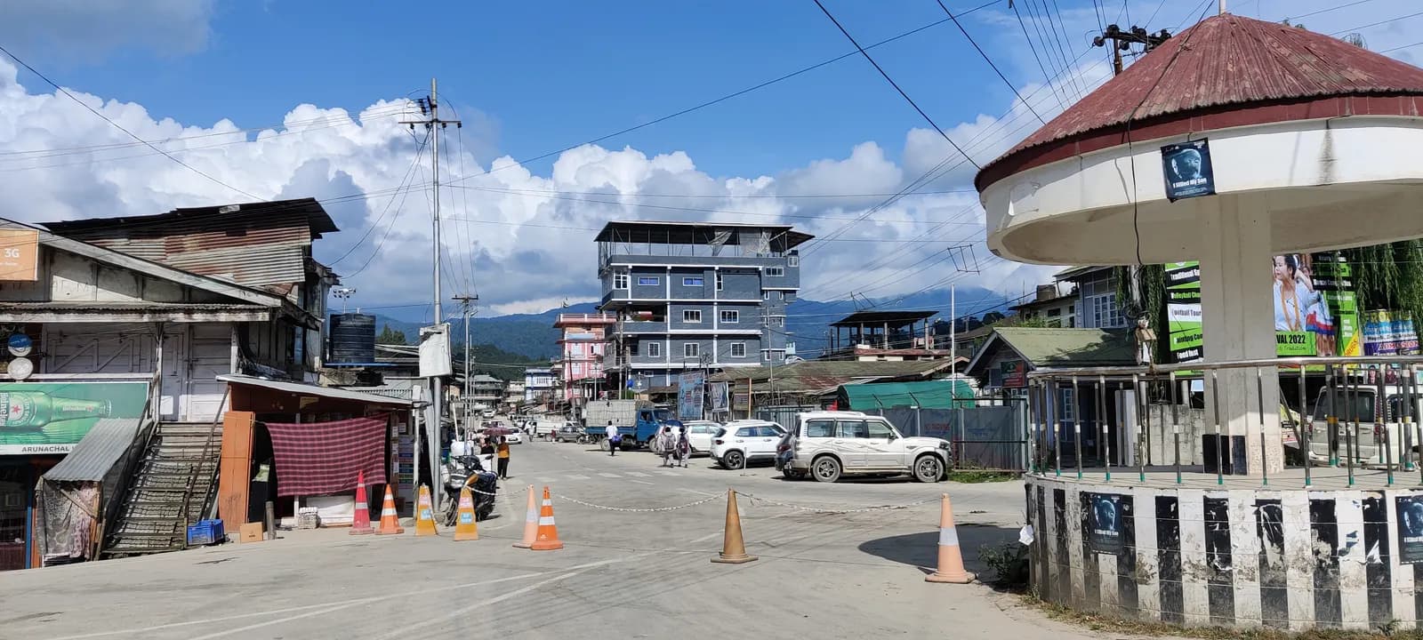 A street junction in Itanagar town with small shops, parked cars and a multi-storey building against the hills