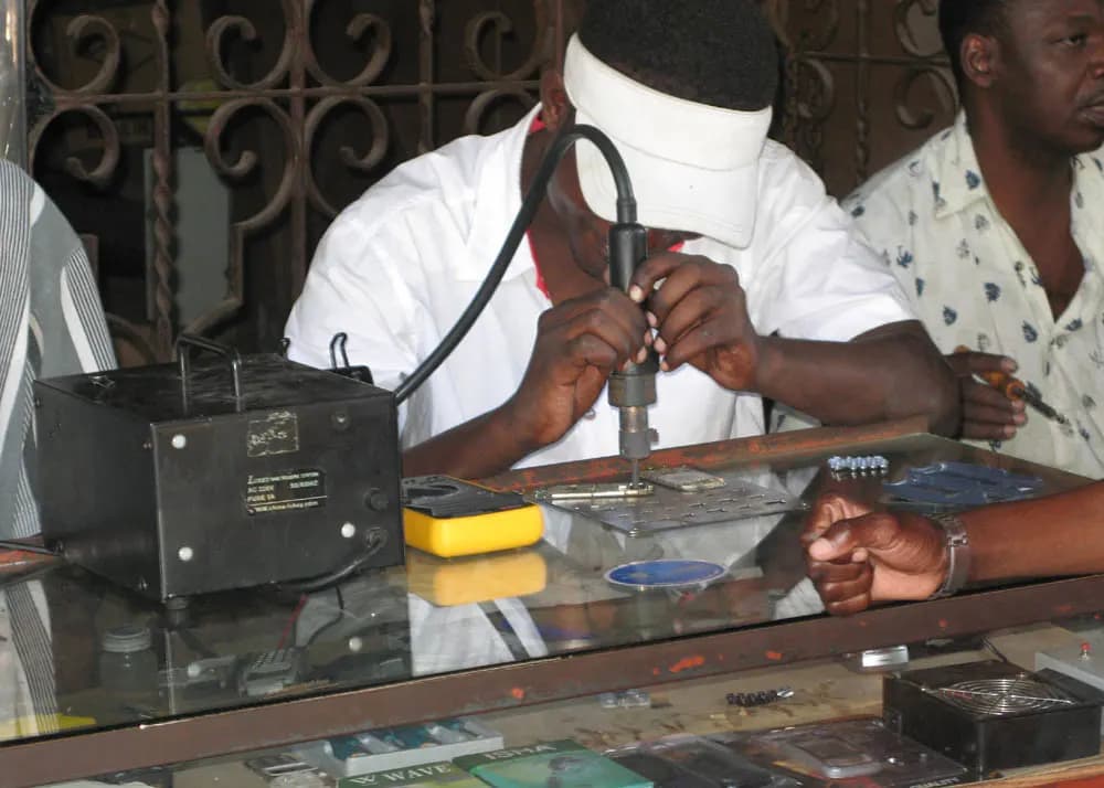 A technician at a glass-topped counter using a hot-air rework pen on a phone board, with a rework station and multimeter beside him