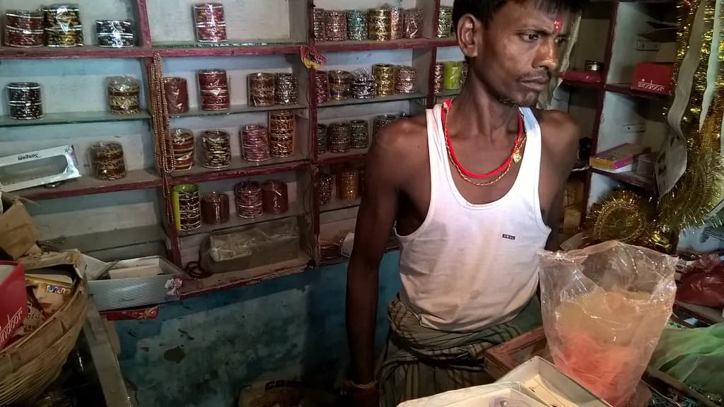 A shopkeeper standing in a small bangle shop in India with glass shelves of bangle boxes behind him