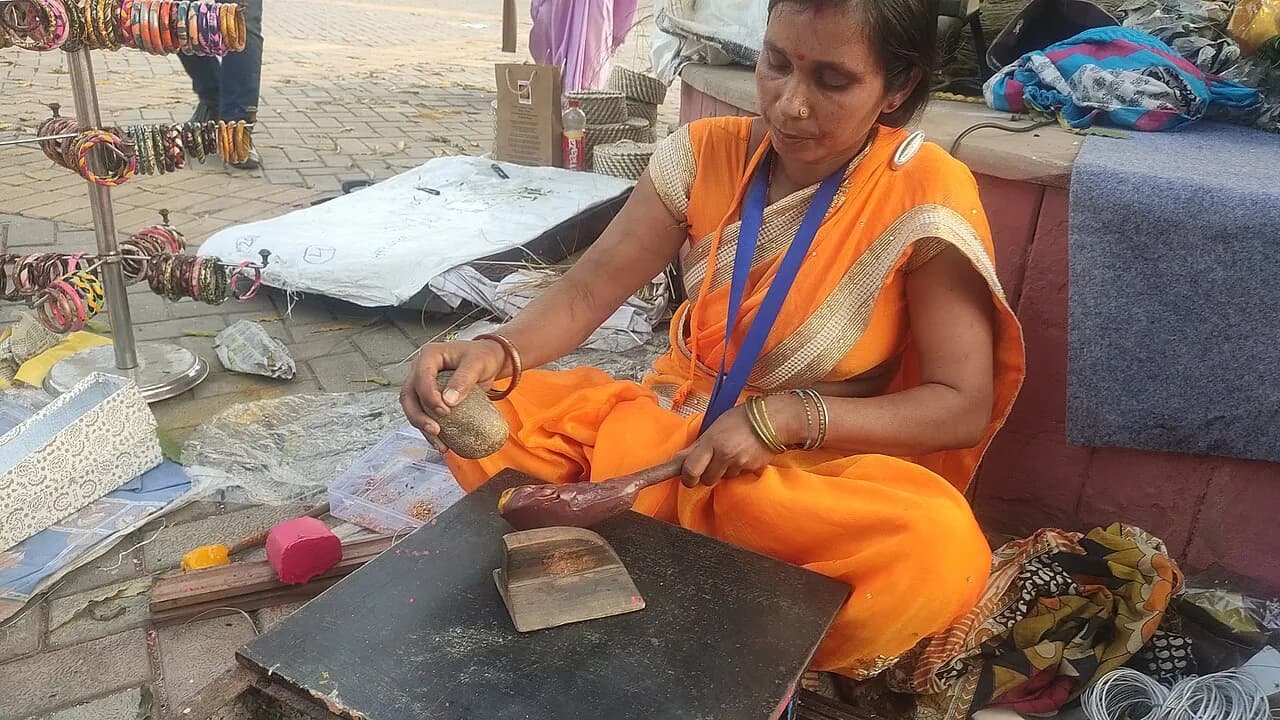 An artisan shaping heated lac on a wooden board to make bangles at a craft fair stall, with finished bangles on a stand