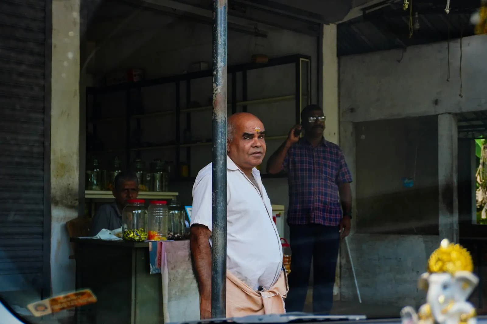 A shopkeeper in a white shirt and mundu standing at the front of a small provisions store in India