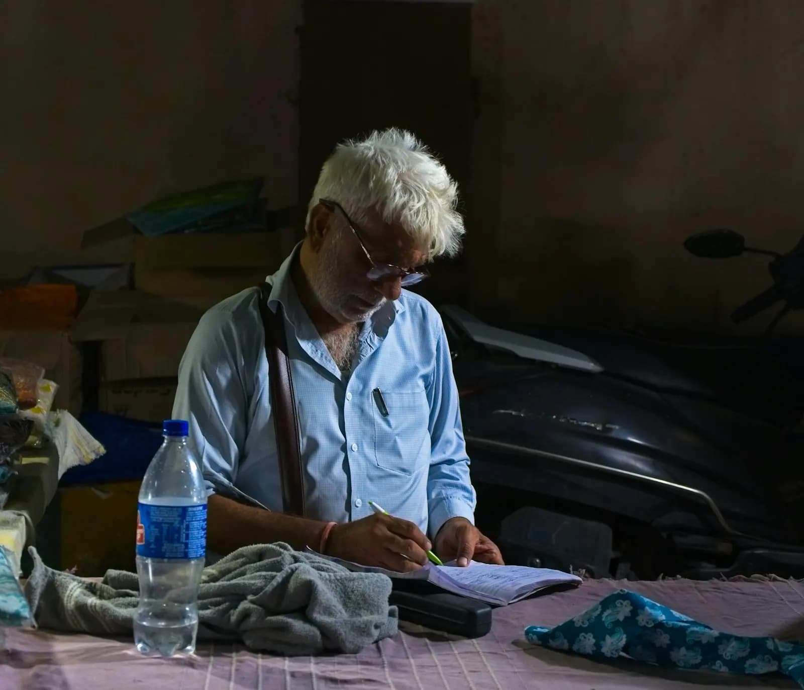 An elderly businessman writing in a notebook at a desk in a small workshop-store