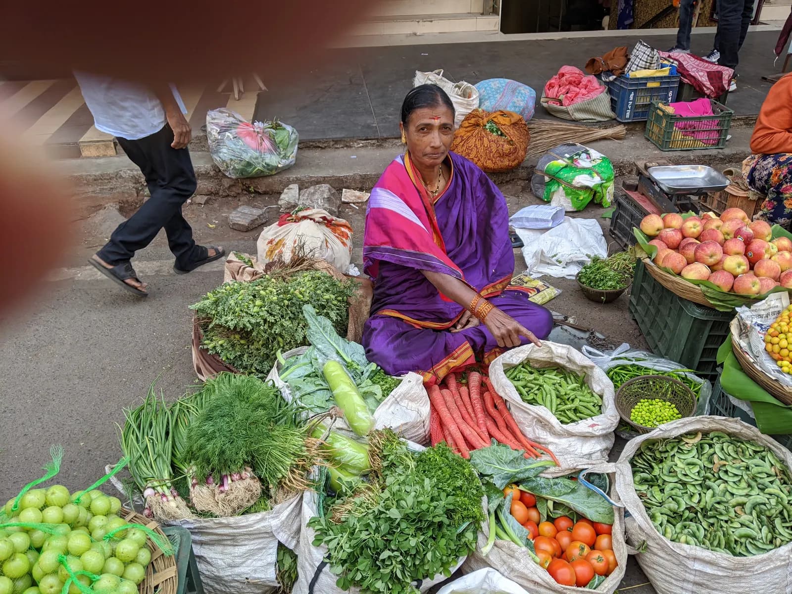 A woman street vendor in a purple sari selling vegetables and fruit from baskets on a pavement in Pune