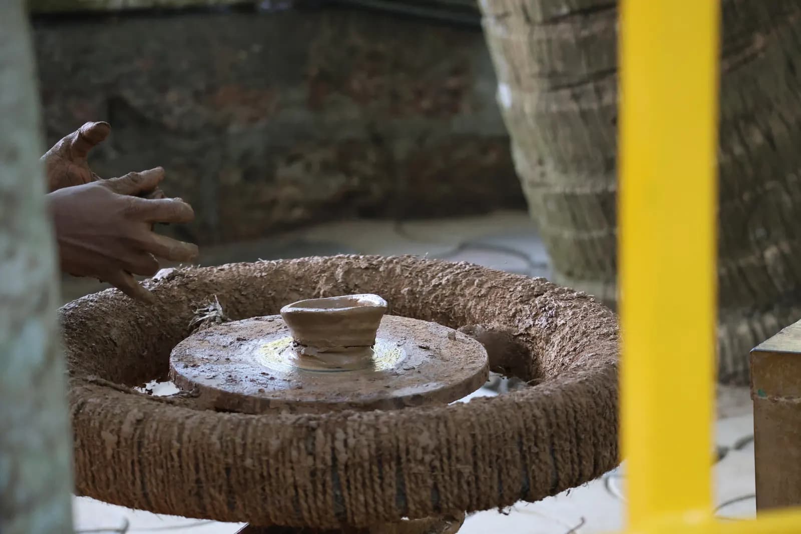 A potter's clay-covered hands beside a small pot on a traditional manual potter's wheel in India