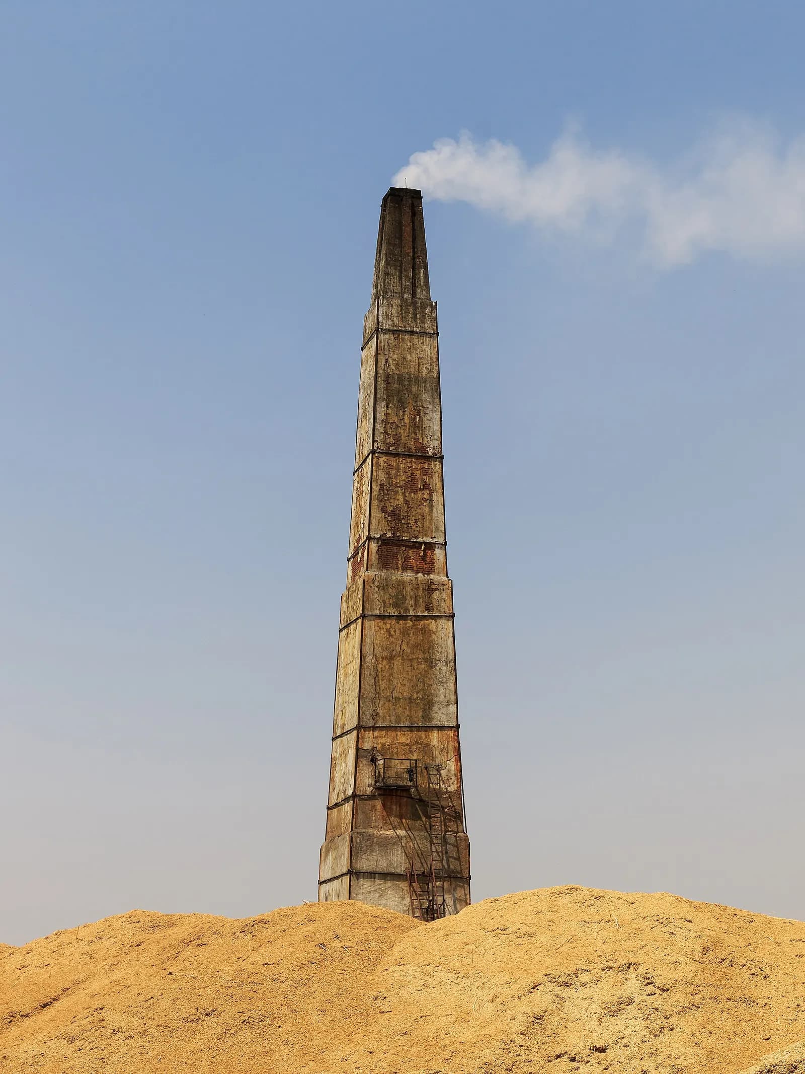 The tall brick chimney of a brick kiln releasing smoke, with a mound of earth in front, in Bharatpur district, Rajasthan