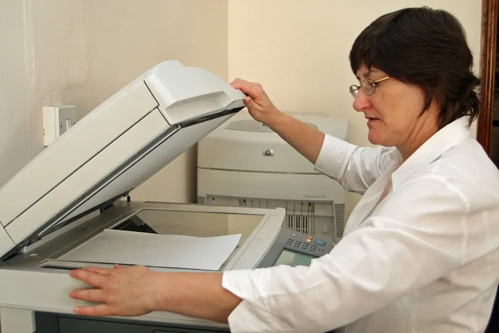An operator lifting the lid of a multifunction photocopier to place a document on the glass, with a laser printer behind