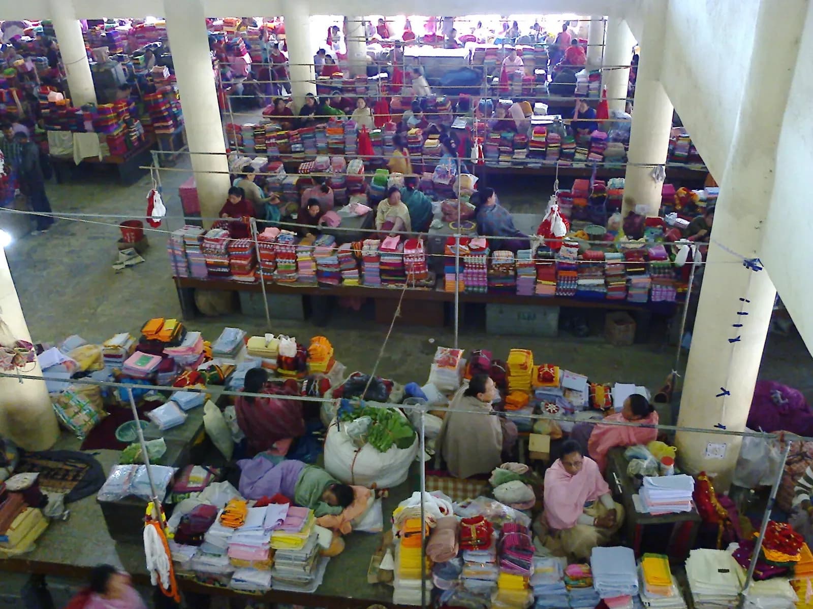 Women traders seated among stalls stacked with cloth and goods in a covered market hall in Imphal, Manipur