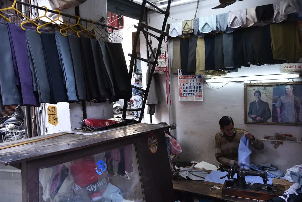 A tailor stitching at his workbench in a small basement shop in Amritsar, with trousers hanging overhead