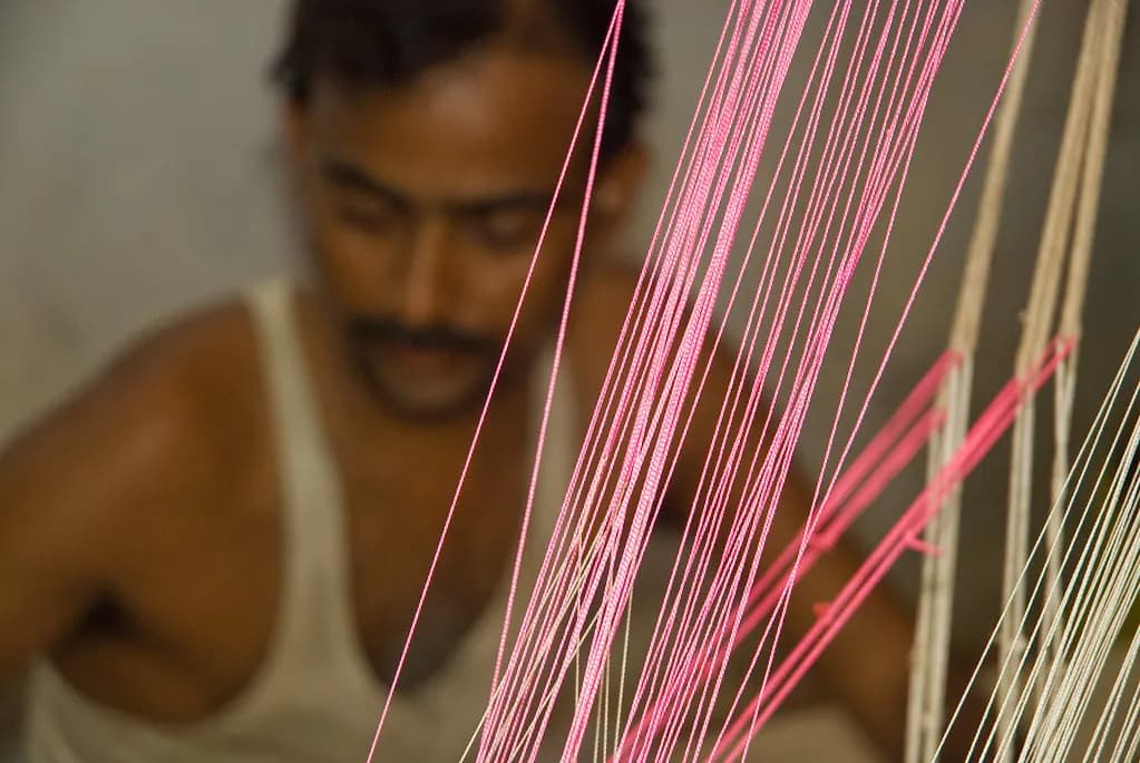 Pink and white warp threads on a loom in a small weaving unit, with the weaver out of focus behind