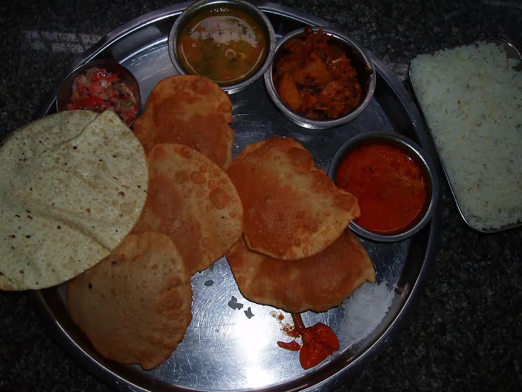 A steel thali with puris, papad, small bowls of curry and a side of rice