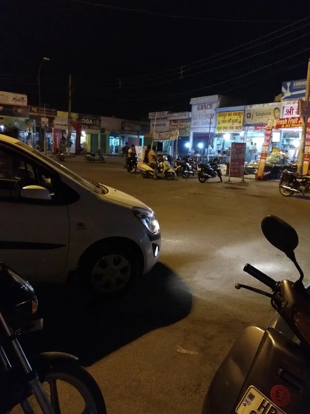 Shops lit up at night in the Sector-1 market, Rohtak, with cars and scooters parked outside