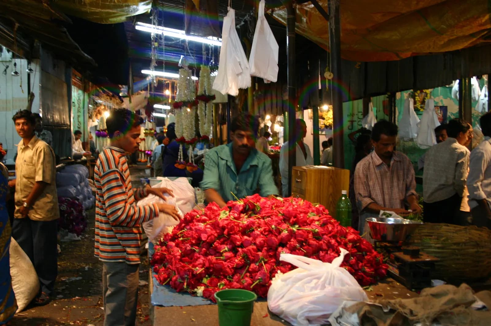 Flower sellers at a stall heaped with red roses inside Devaraja Market, Mysuru, under strip lights