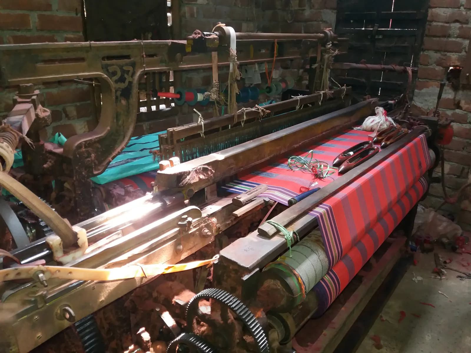 An old shuttle power loom weaving striped red cotton fabric in a brick-walled shed in Hooghly district, West Bengal