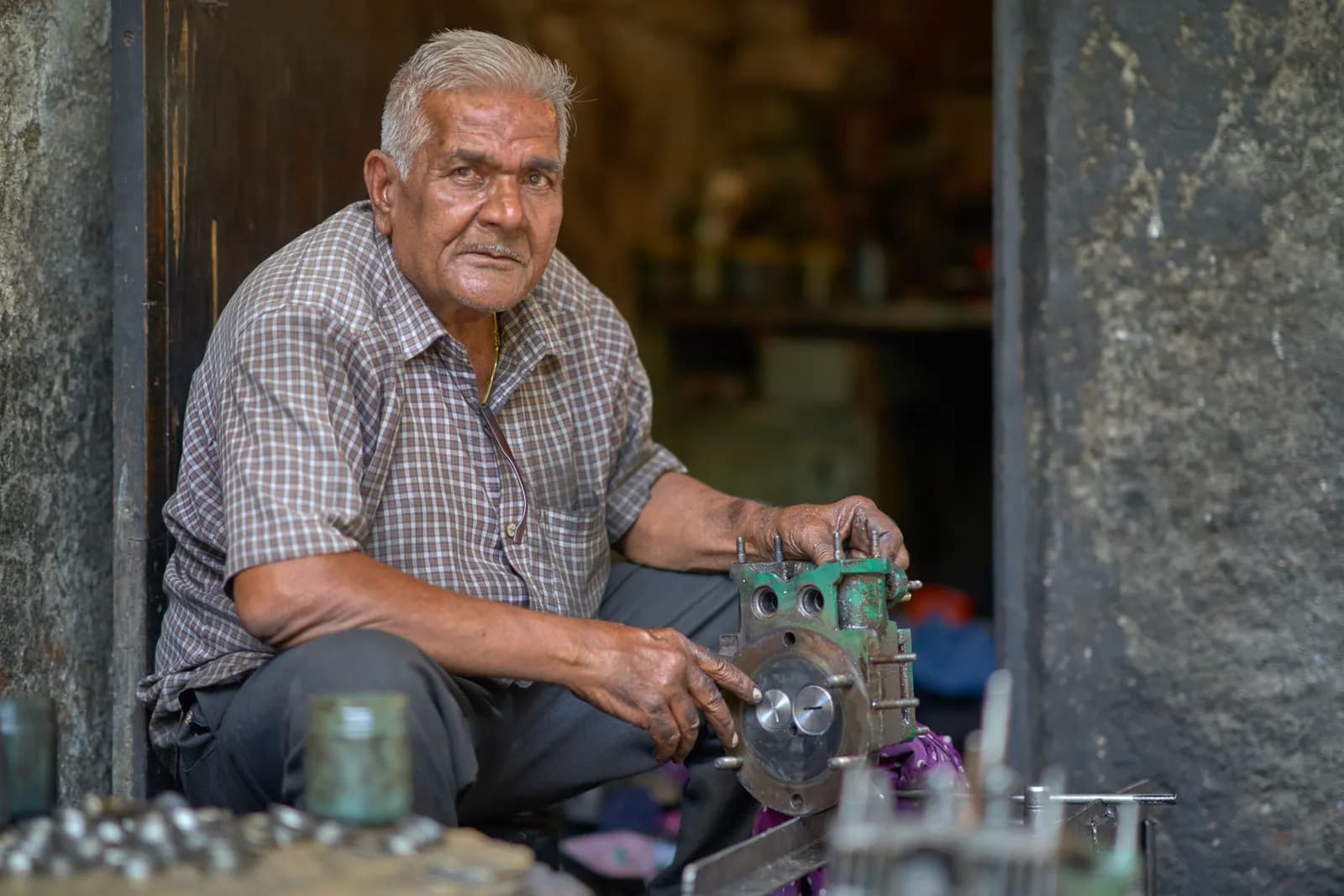 An elderly mechanic in Mumbai sitting at his workshop door with an engine part he is reconditioning