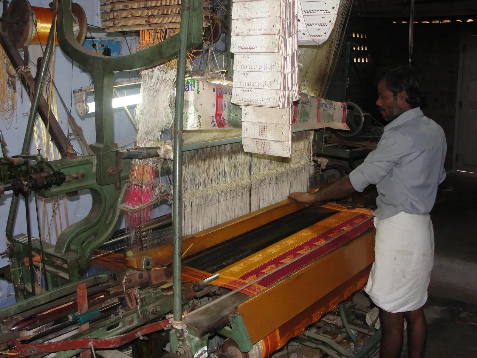 A weaver standing beside a power loom producing a patterned sari border in a small Tamil Nadu weaving unit