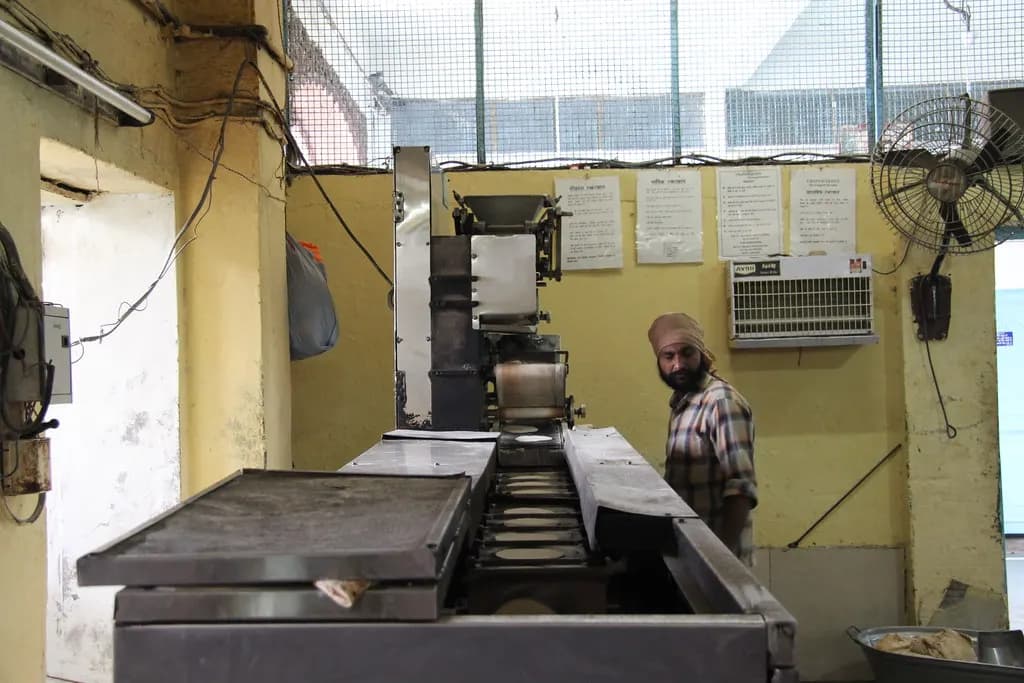 A stainless steel automatic chapati-making machine with a conveyor and griddle in a large community kitchen