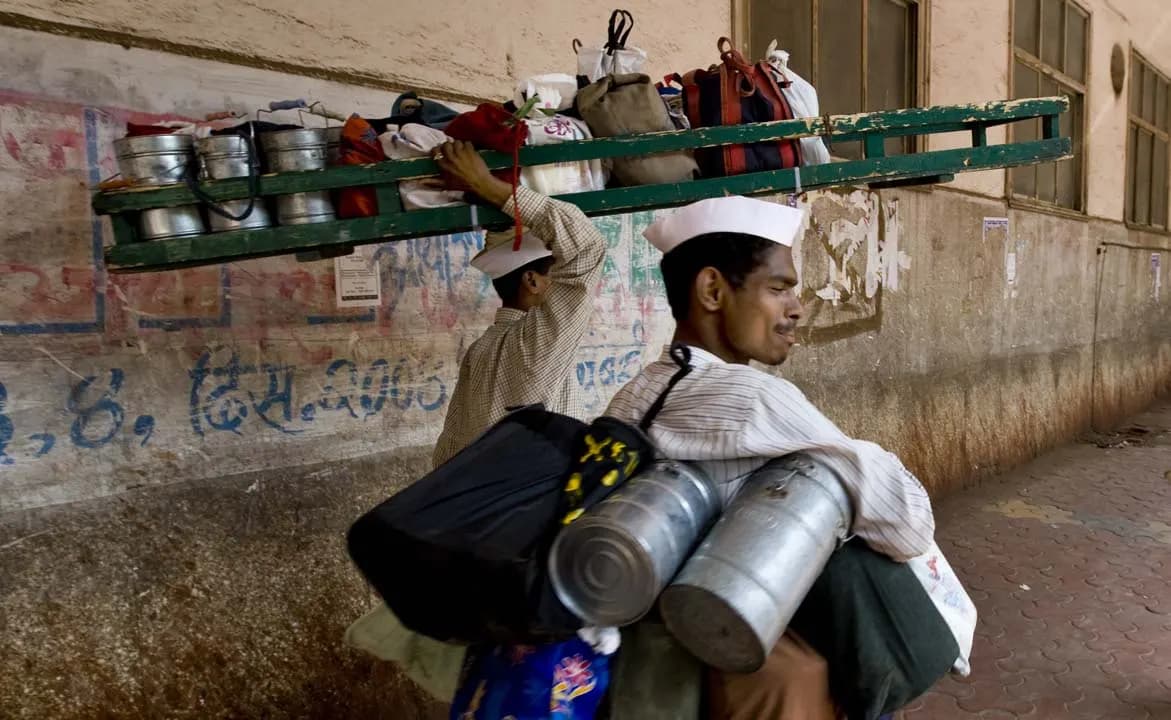 Two Mumbai dabbawalas carrying a long wooden crate of steel tiffin carriers and cylindrical lunch boxes along a street