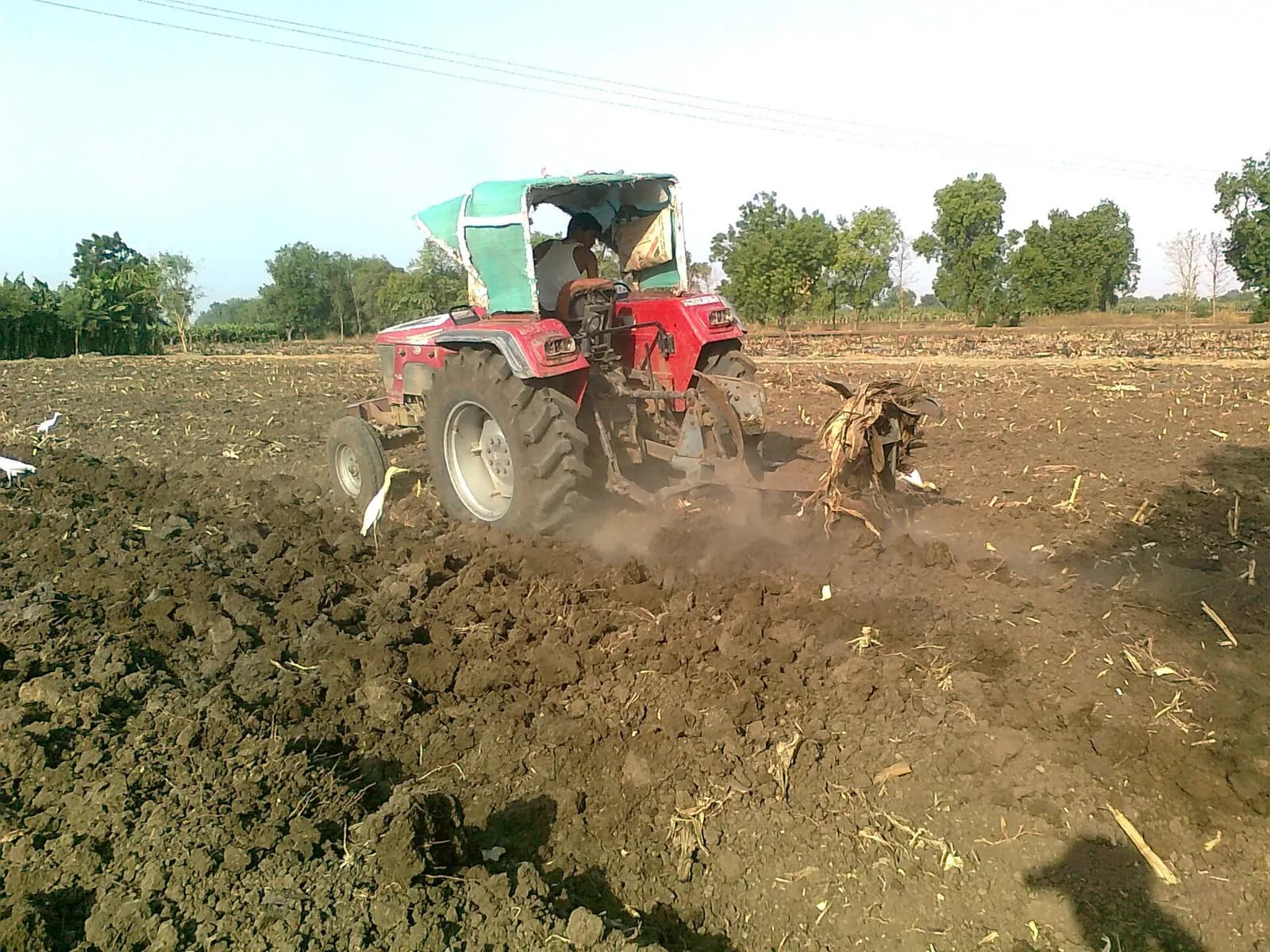 A farmer ploughing a dry field with a red tractor and mounted plough in rural India