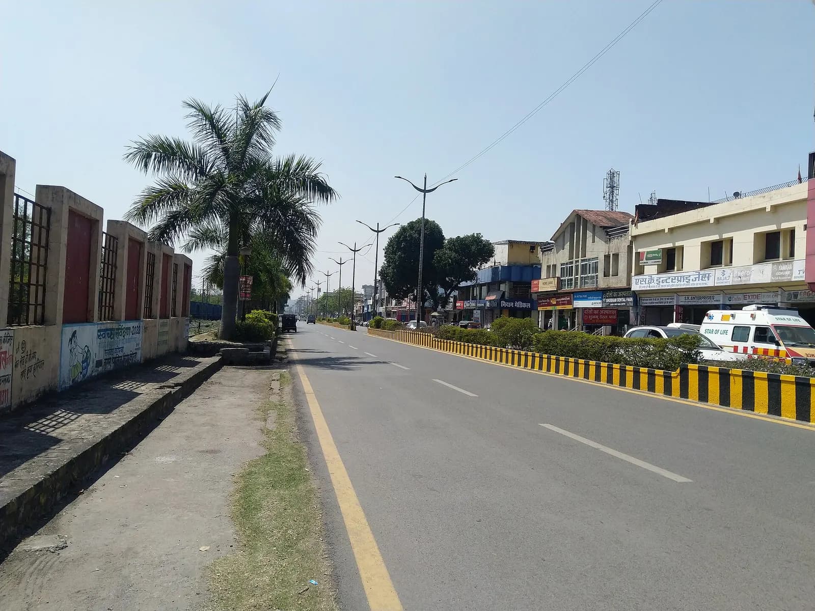 A wide divided road in TP Nagar, Korba, with a row of two-storey shops and offices along one side