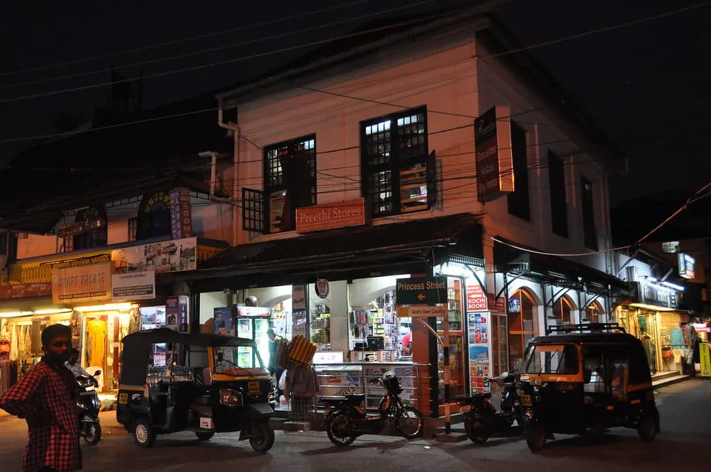 Shopfronts and autorickshaws on Princess Street in Fort Kochi at night, with a small general store lit up on the ground floor