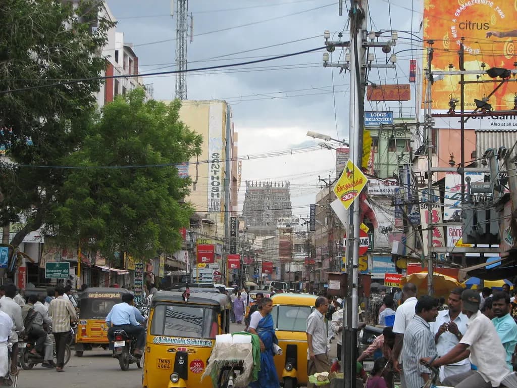 A busy commercial street in Madurai with shops, auto-rickshaws and the Meenakshi temple tower in the distance