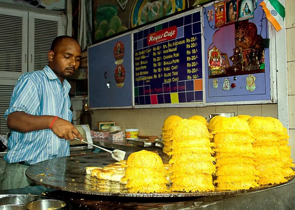 A cook frying aloo tikki at the counter of a chaat eatery in Hazratganj, Lucknow, with baskets of fried basket chaat stacked beside him