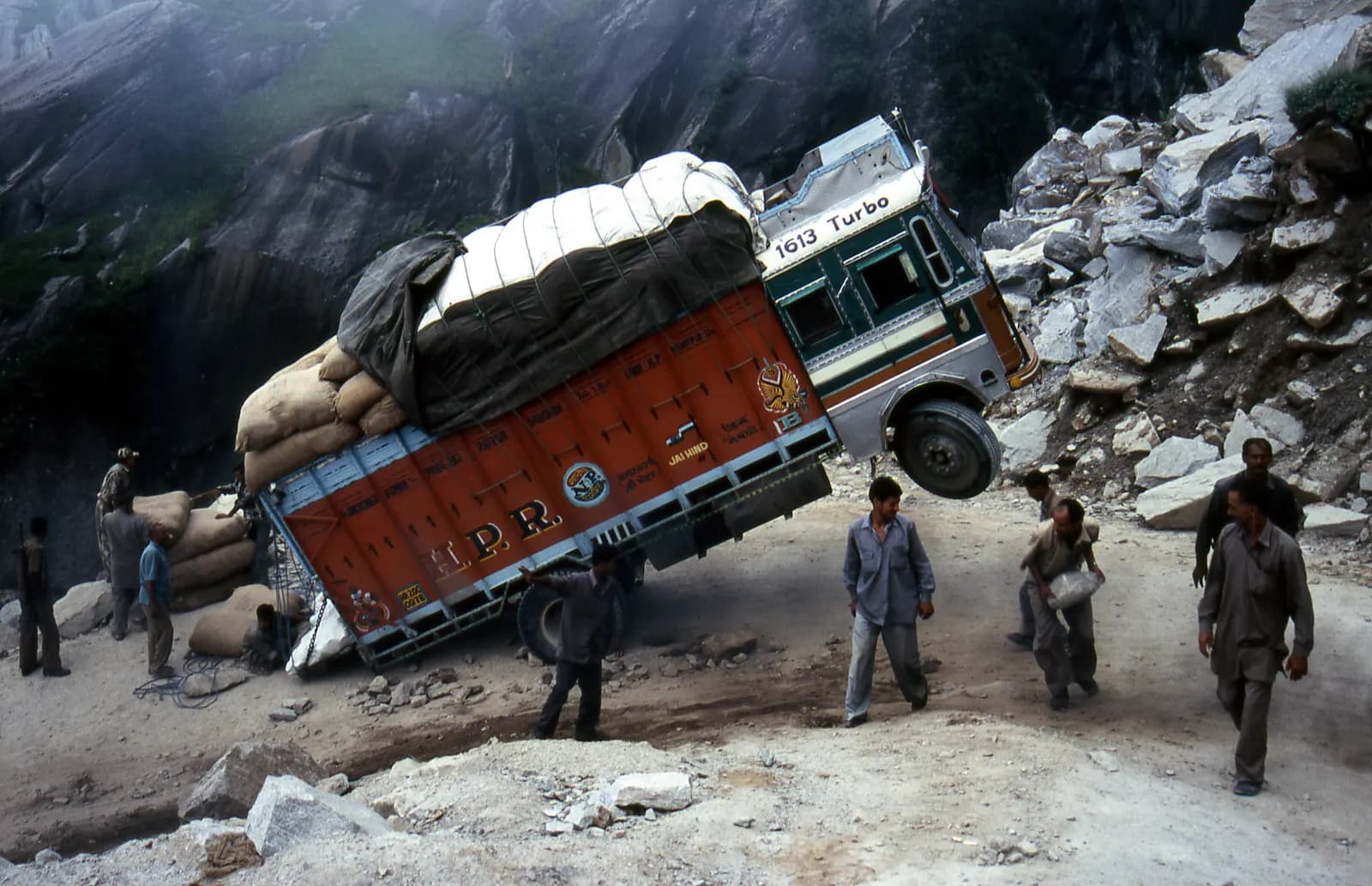 An overloaded Indian goods truck tilted on a rocky mountain road while people stand around it