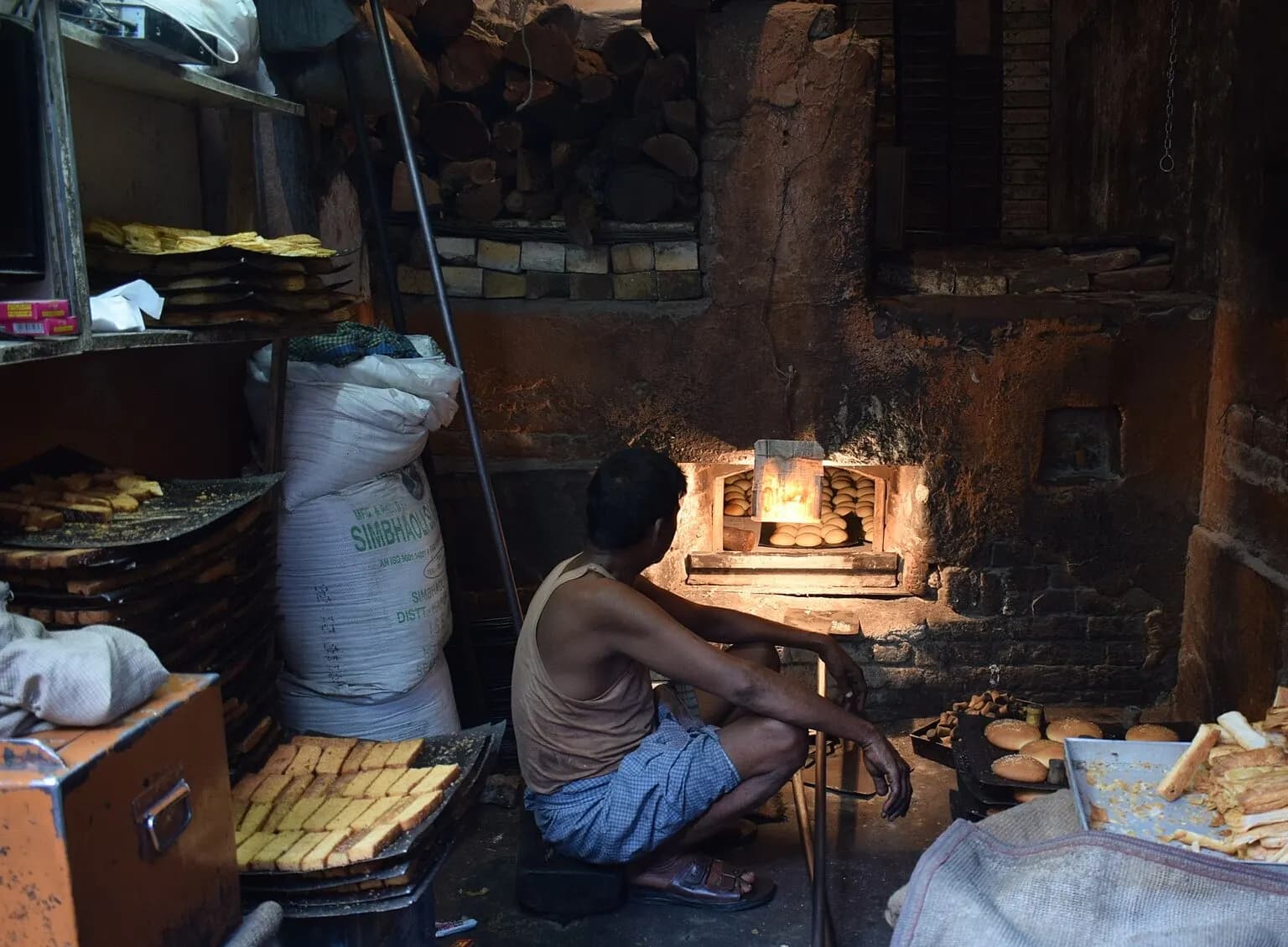 A worker tending a wood-fired oven in a small Indian bakery, with trays of rusk and sacks of flour around him