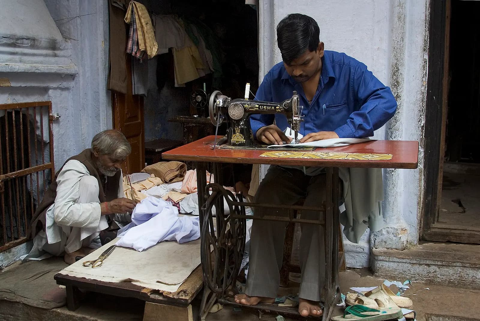 A tailor stitching at a treadle sewing machine while an older craftsman cuts cloth beside him in a Varanasi street workshop