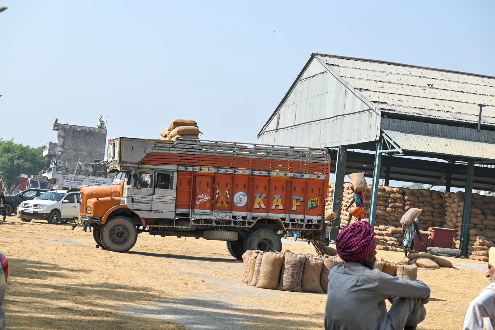 Sacks of grain stacked under an open shed at a grain market yard in north India, with a loaded truck and a farmer waiting