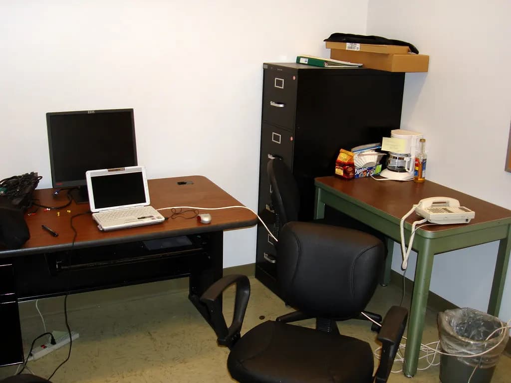 A small office with a desk, laptop, monitor, desk phone and a black filing cabinet