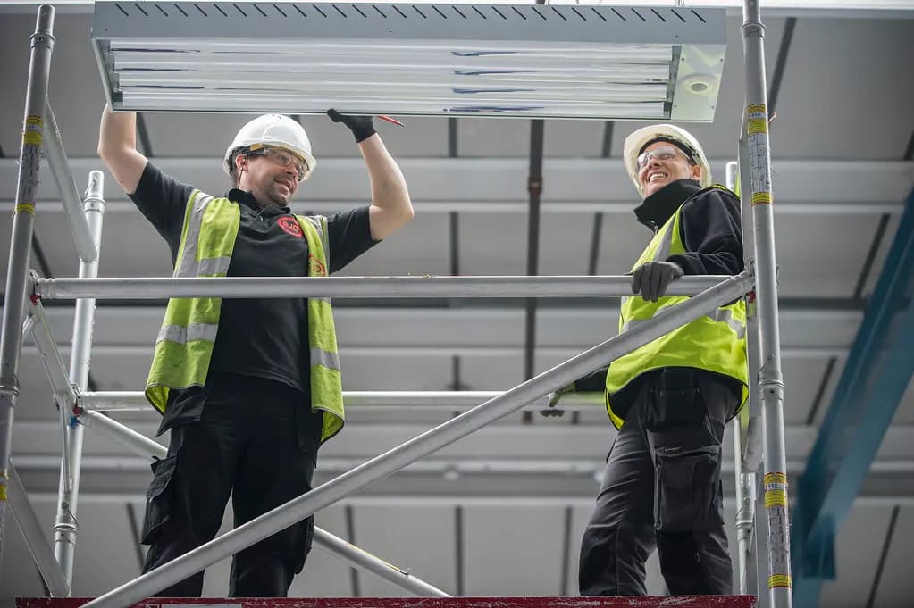 Two electricians in hard hats and hi-vis vests fitting ceiling lights from a mobile scaffold tower