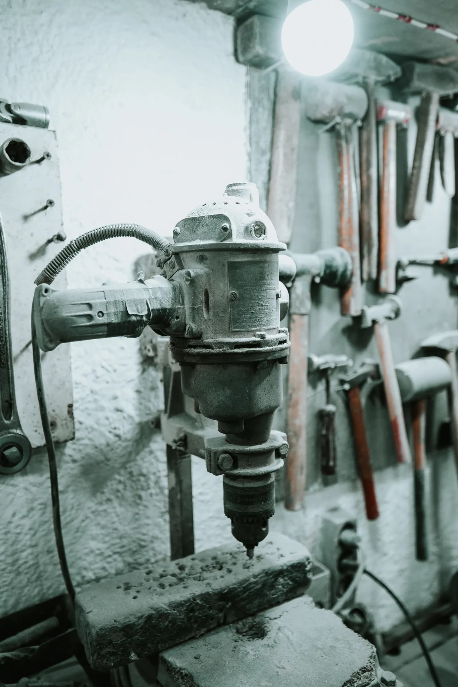 A dusty drill mounted in a stand on a workbench, with hammers hanging on the workshop wall behind