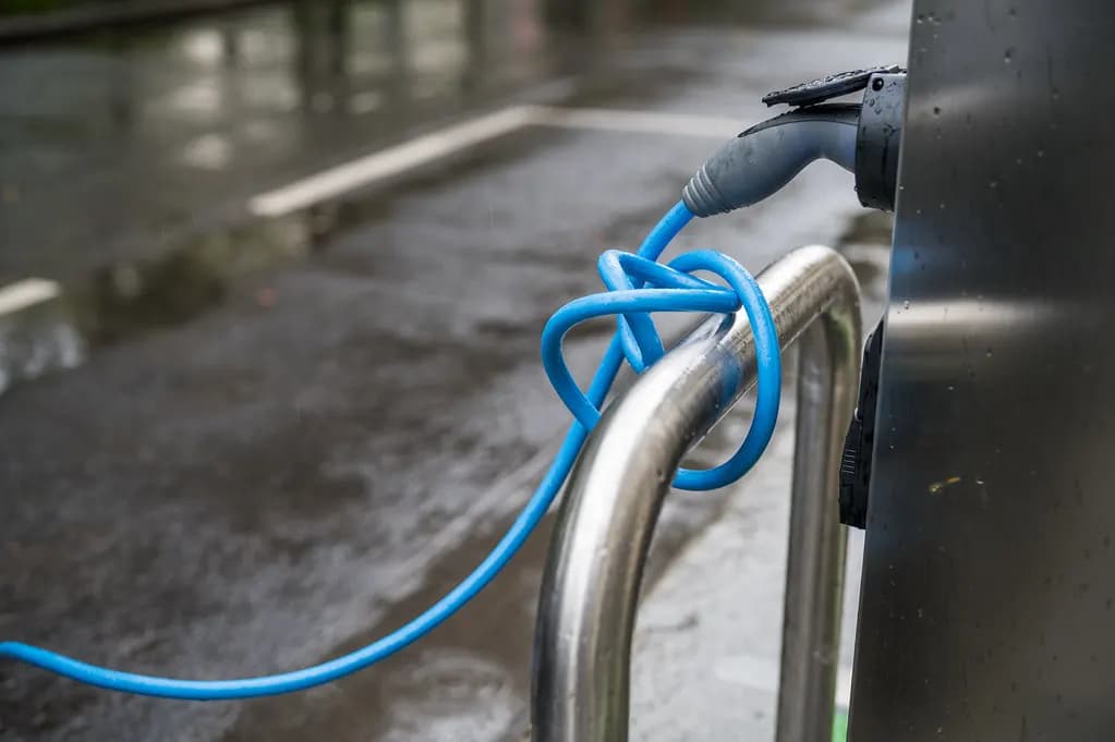 A blue charging cable plugged into an electric vehicle charging post on a wet street