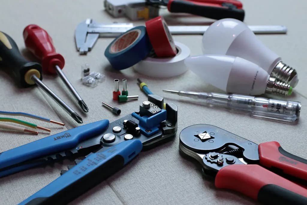 Electrician's hand tools laid out on a table: wire strippers, crimping pliers, screwdrivers, insulation tape, cable and light bulbs