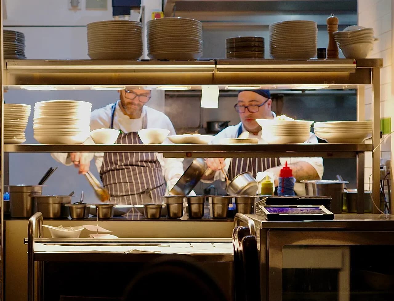 Two chefs in aprons cooking behind a pass stacked with plates in a UK restaurant kitchen
