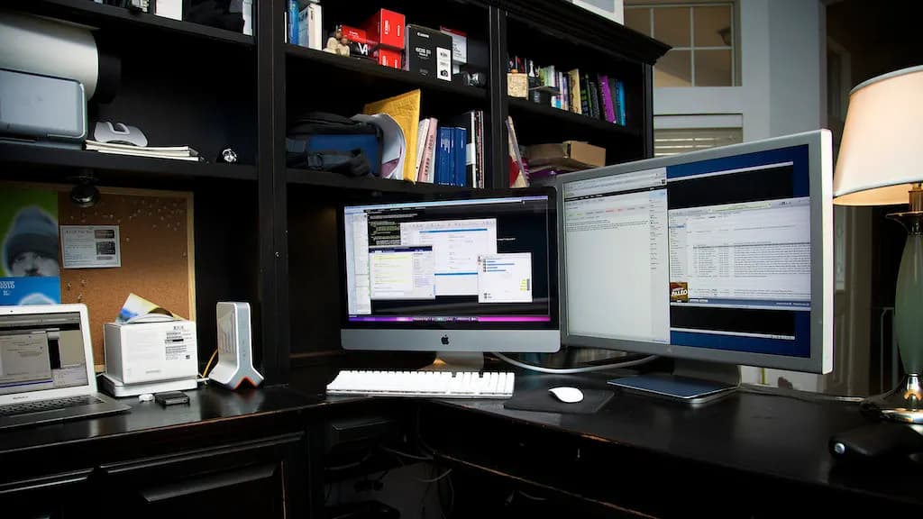 A home office desk with a large monitor, laptop, keyboard and desk lamp
