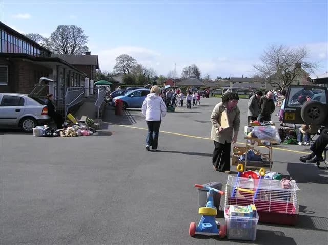 Household items, toys and a pet cage laid out for sale beside parked cars at a car boot sale in a school car park
