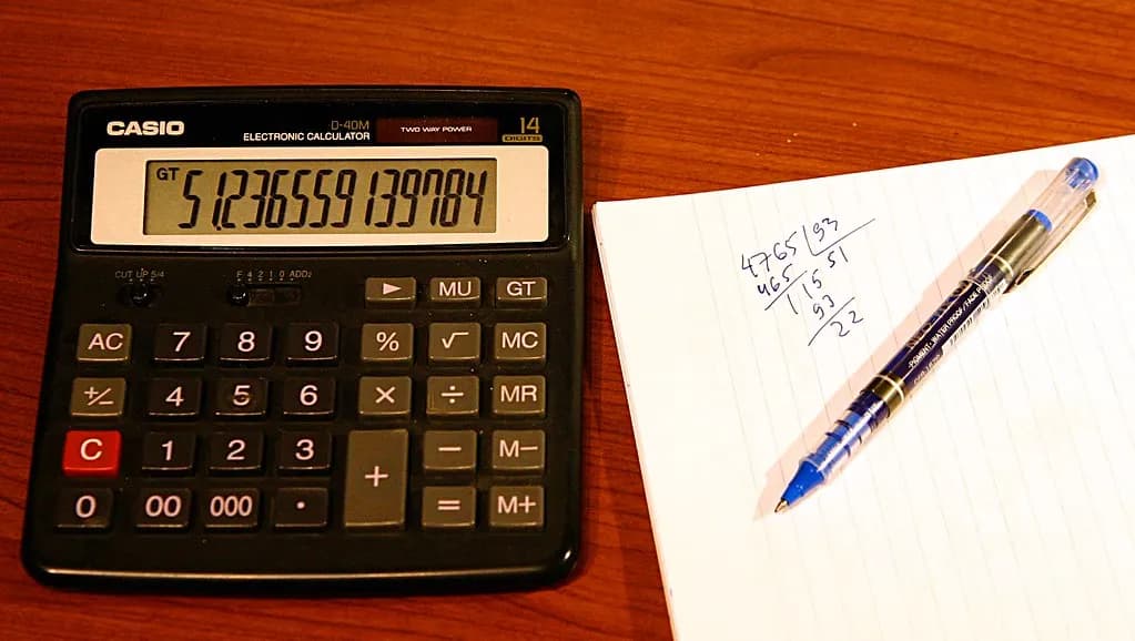 A desk calculator next to a notepad with handwritten sums and a ballpoint pen