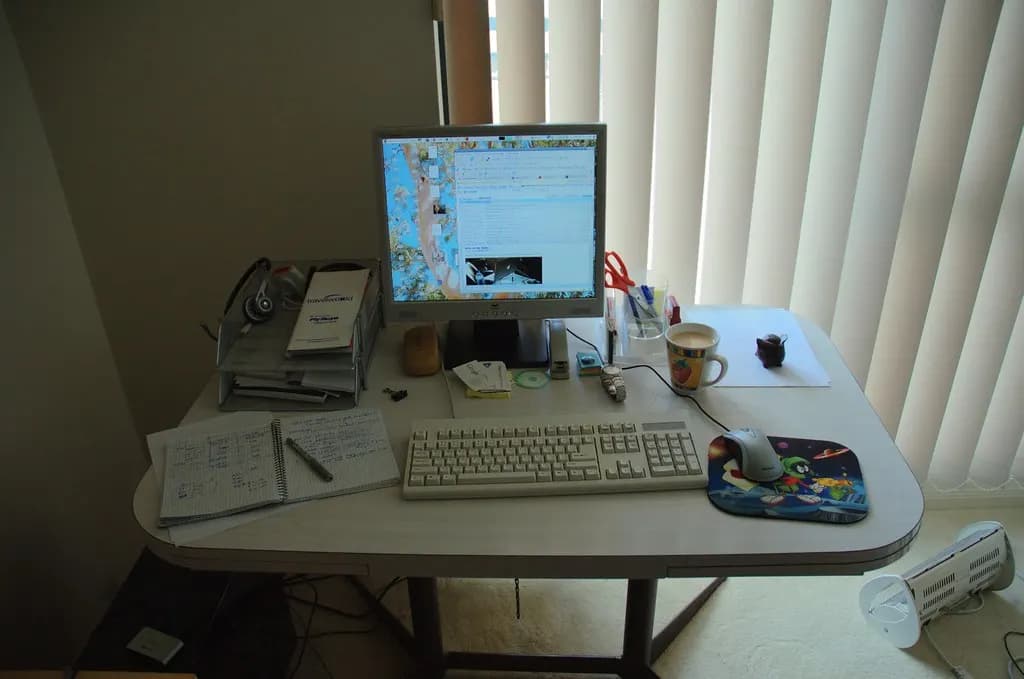 A home working area with a desk, computer screen, keyboard and paperwork by a window