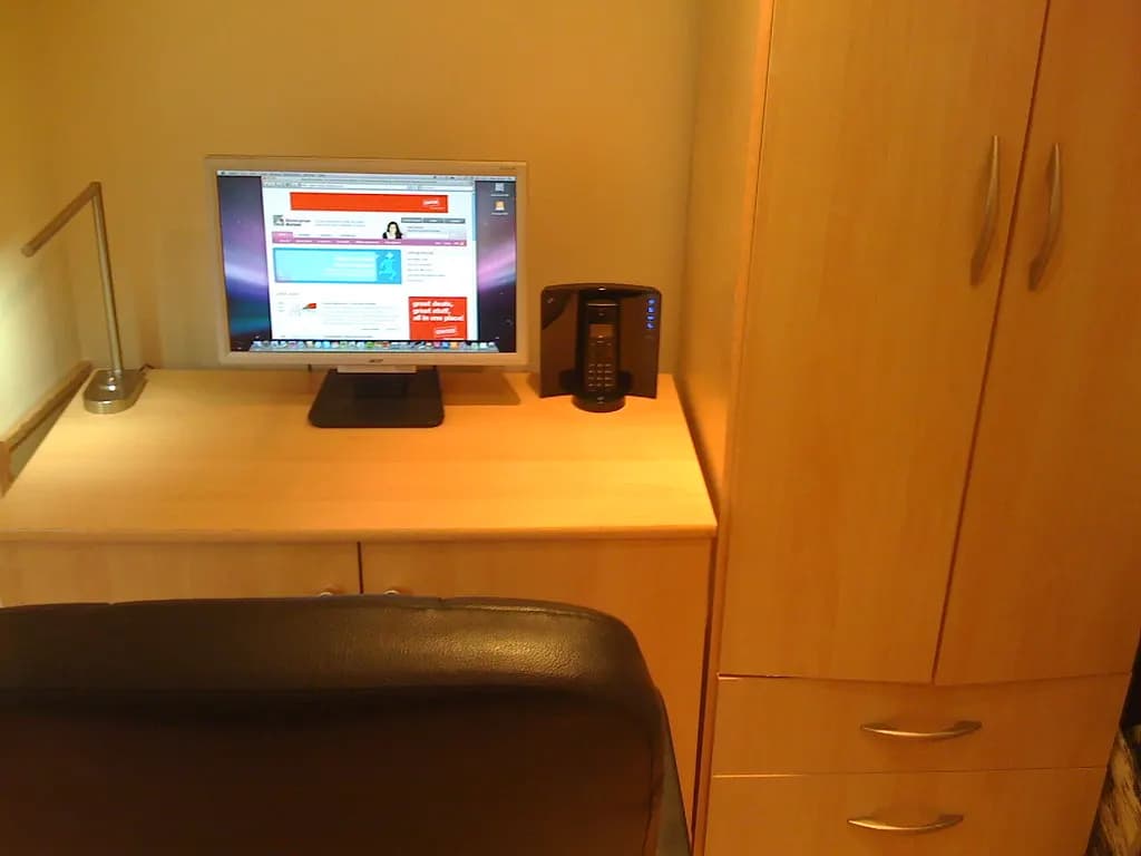 A desk with a computer screen, lamp and phone set beside a wardrobe in a small room of a house