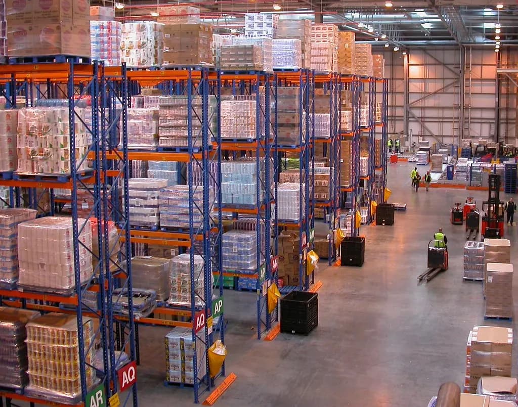 Distribution centre with tall pallet racking stacked with cartons of household goods and staff moving pallets on electric trucks